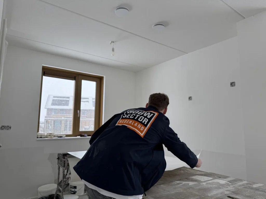 Behanger in donkere werkjas met logo pakt een baan behang van de plaktafel in een lege nieuwbouwkamer met besneeuwde huizen buiten