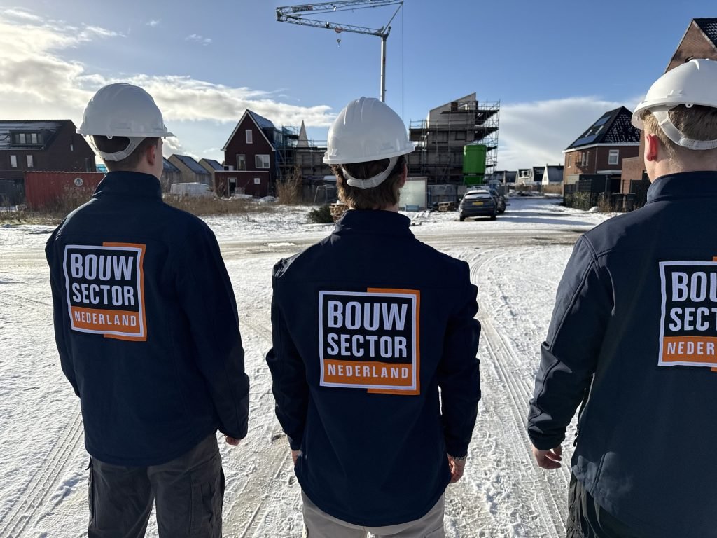 Drie vakmensen van Bouwsector Nederland met witte bouwhelm op een besneeuwde bouwweg tussen nieuwe woningen en een kraan